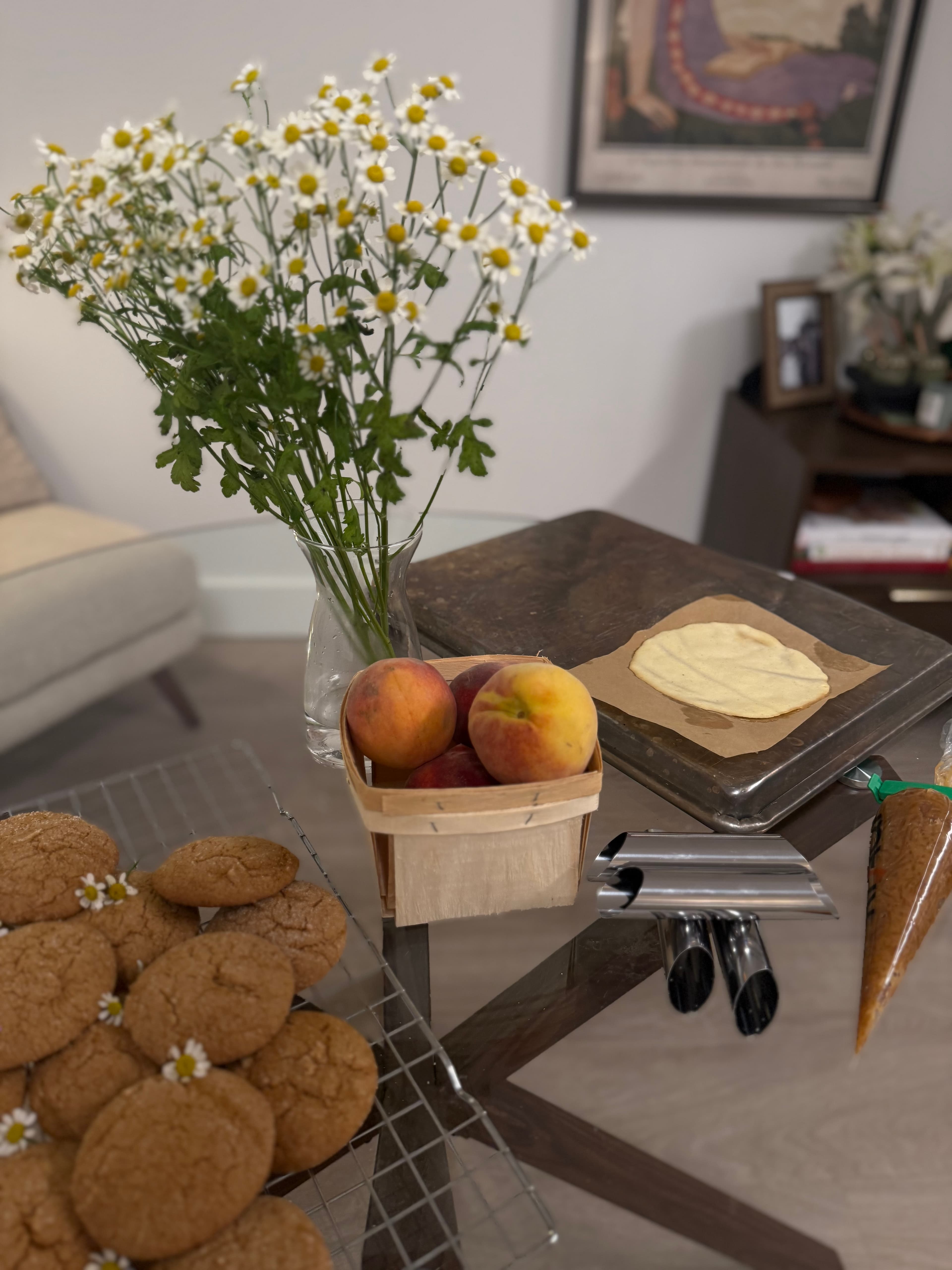Fresh-baked cookies cooling on a rack, alongside chamomile blooms, peaches, and a rolled dough round mid-prep